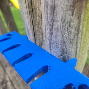 May include: A close-up of a blue plastic tool with multiple slots. The tool is attached to a wooden post.