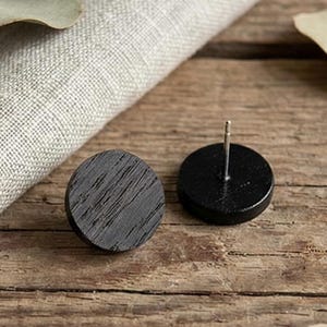 May include: Two black wooden stud earrings. One earring is face up, showing the wood grain, and the other is face down, showing the post. The earrings are on a wooden surface with a linen cloth and dried leaves.