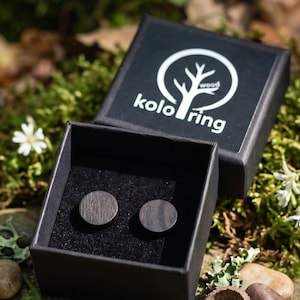 May include: A pair of dark wood stud earrings presented in a black box, branded with the "kolo ring wood" logo. The box is open, displaying the earrings on black fabric. The background features green moss, small white flowers, and natural elements.