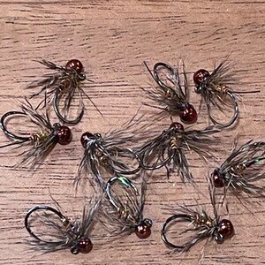 May include: A close-up of nine brown and black fly fishing lures with brown beads. The lures are arranged on a wooden surface.