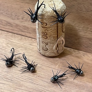 May include: Five black fly fishing lures with iridescent green and black bodies and long, thin, black legs. The lures are arranged on a wooden surface and a cork.