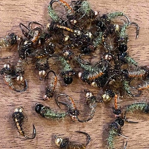 May include: A close-up of a pile of small, brown and green fishing flies with black beads. The flies are tied with brown and gold thread and have a variety of colors and patterns.