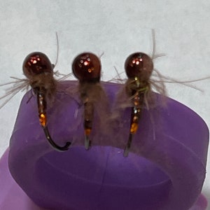 May include: Three brown and gold fly fishing lures with brown feathers and a purple ring.
