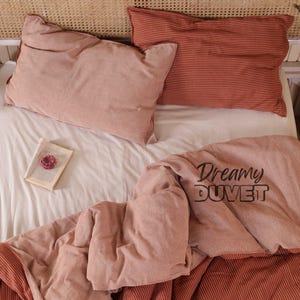 May include: A bed set featuring a light pink duvet cover and pillowcases. The duvet cover has the text "Dreamy DUVET". One pillowcase is plain pink, the other is striped. A book with a pink flower rests on the white sheet.