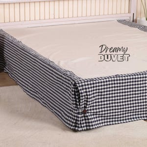 Puede incluir: Una cama con cabecero blanco y colchón color crema. La falda de la cama es a cuadros azul marino y blanco. El texto "Dreamy DUVET" está impreso en el colchón.