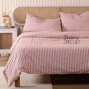 May include: A bed dressed with a duvet cover and two pillowcases. The bedding has a striped design in red and white. The words "Dreamy DUVET" are printed on the duvet cover. The headboard is made of woven rattan.