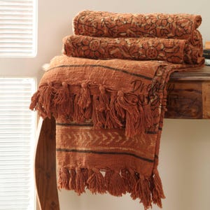 Puede incluir: Una manta de color óxido con un patrón de rayas y ribete de flecos. La manta está doblada sobre una mesa de madera.