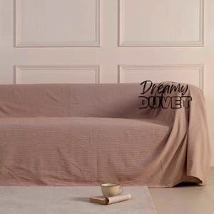 Könnte beinhalten: Ein rosafarbenes Sofa ist mit einem gestreiften Überwurf bedeckt. Der Überwurf hat dünne, dunkle Streifen. Vor dem Sofa liegt ein kleiner, cremefarbener Teppich. Eine Tasse und ein Buch liegen auf dem Teppich. Der Text "Dreamy Duvet" ist auf dem Überwurf.