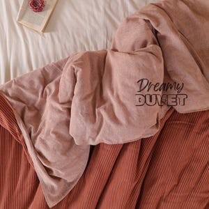 May include: A bed featuring a white sheet, a pink duvet, and a rust-coloured striped blanket. An open book with a pink flower rests on the bed. The text "Dreamy DUVET" is printed on the duvet.