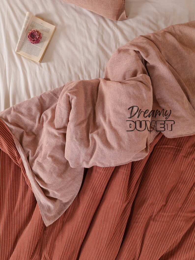 May include: A bed featuring a white sheet, a pink duvet, and a rust-coloured striped blanket. An open book with a pink flower rests on the bed. The text "Dreamy DUVET" is printed on the duvet.
