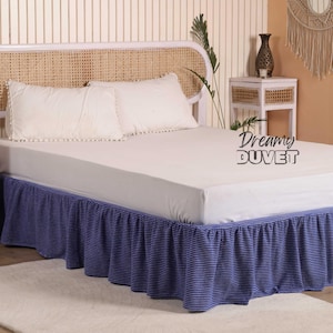 Puede incluir: Una cama con una sábana bajera blanca y dos almohadas blancas con ribete de pompón. La falda de la cama es azul con finas rayas blancas. El cabecero es blanco con un diseño tejido. El texto "Dreamy DUVET" es visible.