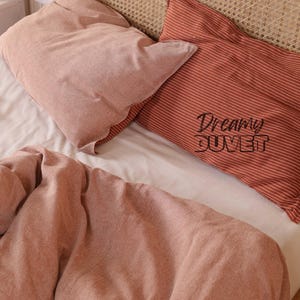 May include: A bed with a white sheet, two pillows, and a duvet. One pillow is solid pink, and the other is red with white stripes. The red pillow has the words "Dreamy DUVET" printed on it. The duvet is pink.