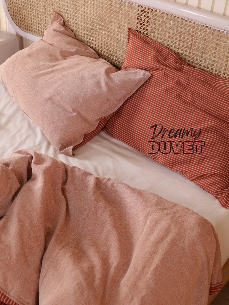 May include: A bed with a white sheet, two pillows, and a duvet. One pillow is solid pink, and the other is red with white stripes. The red pillow has the words "Dreamy DUVET" printed on it. The duvet is pink.