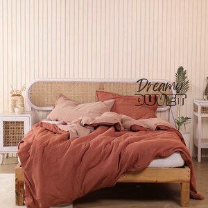 May include: A bed with a wooden frame and a woven rattan headboard. The bedding features a rust-coloured striped duvet cover, pillows, and a light pink sheet. The text "Dream Duvet" is visible on the headboard.