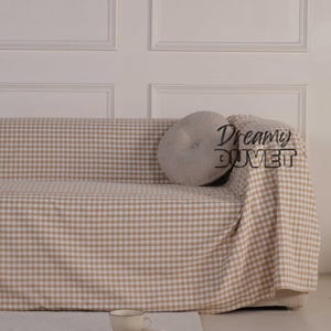 May include: A beige and white checkered sofa cover draped over a sofa, with a matching pillow and blanket. A cup and a book rest on a white rug in front of the sofa. The text "Dreamy Duvet" is visible on the blanket.