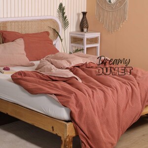 May include: A bed with a wooden frame and a woven headboard. The bedding features a red-orange striped duvet, matching pillowcases, and a light pink pillow. The text "Dreamy DUVET" is displayed on the duvet.