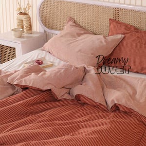 May include: A bed with a white headboard and a woven cane insert. The bedding includes a duvet and two pillows in shades of pink and red. The duvet cover has a striped pattern. The text "Dreamy DUVET" is visible.