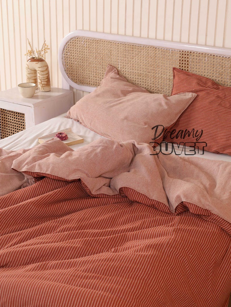 May include: A bed with a white headboard and a woven cane insert. The bedding includes a duvet and two pillows in shades of pink and red. The duvet cover has a striped pattern. The text "Dreamy DUVET" is visible.