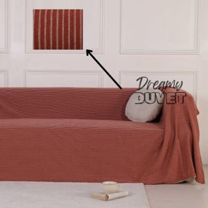 May include: A terracotta-coloured sofa cover with vertical stripes, draped over a sofa. A matching throw blanket is draped over the arm. A small, round, grey pillow and a cup and book are on the sofa.