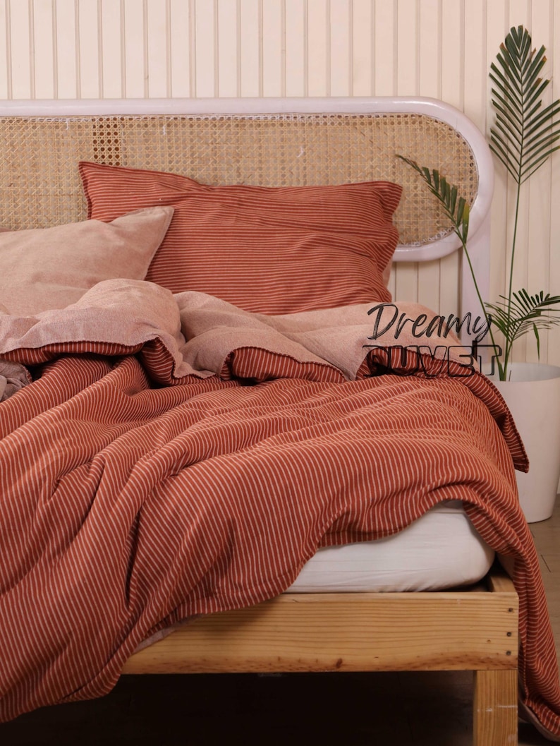 May include: A bed dressed with rust-coloured striped bedding, including a pillow and a duvet cover. The bed frame is made of light-coloured wood, and the headboard has a woven design. A plant in a white pot is visible.