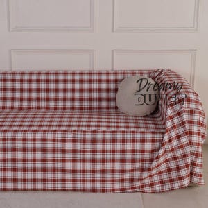 以下が含まれることがあります： 赤と白のチェック柄のソファカバーがかかったソファ。 「Dreamy Dust」という文字が入った丸い灰色のクッションがソファにあります。 カップと本がソファの前の床にあります。