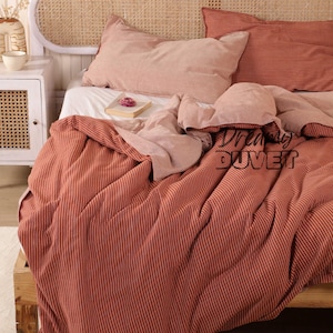 May include: A bed featuring a rust-coloured duvet cover and pillow set with white vertical stripes. The bed has a woven headboard and a white bedside table. The text "Dreaming Duvet" is visible on the duvet cover.