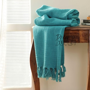 Puede incluir: Una manta de tiro azul turquesa con flecos en el borde inferior. La manta está doblada y drapeada sobre una mesa de madera.