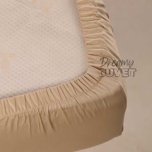Può includere: Un primo piano di un lenzuolo con angoli elastici beige. Il lenzuolo è realizzato in un tessuto morbido e liscio ed è progettato per adattarsi perfettamente a un materasso.