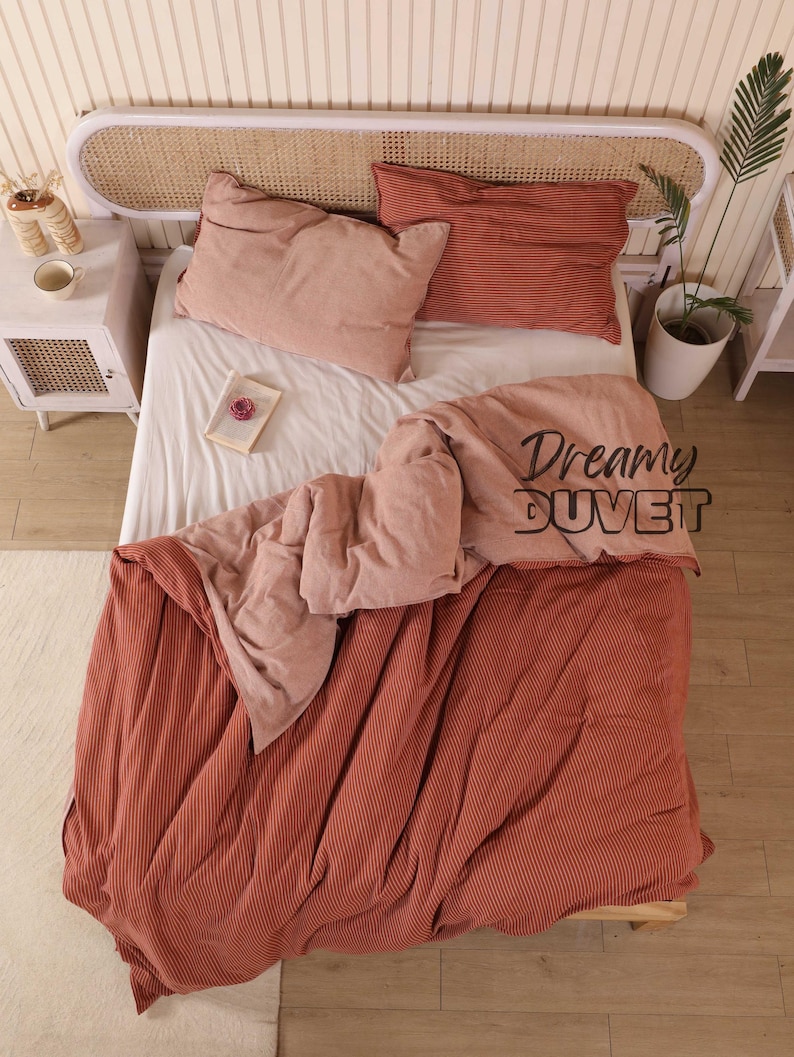 May include: A bed with a white sheet, two pink pillows, and a rust-coloured striped pillow. A rust-coloured duvet with the text "Dreamy DUVET" is draped over the bed. The bed has a rattan headboard and is in a well-lit room.