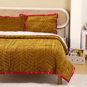 Könnte beinhalten: Ein Bett mit einer senffarbenen Samtsteppdecke und passenden Kissenbezügen. Die Steppdecke und die Kissenbezüge haben ein geometrisches Steppmuster und sind mit einem roten Satinrand versehen. Das Bett hat ein weißes Kopfteil.