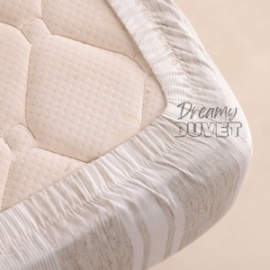 Puede incluir: Primer plano de una sábana bajera con un patrón de rayas blancas y beige. La sábana está hecha de un tejido suave y transpirable y está diseñada para ajustarse perfectamente a un colchón.