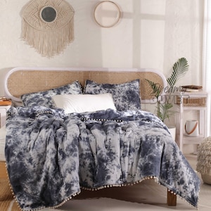 May include: A blue and white tie-dye duvet cover with pom pom trim. The duvet cover is on a bed with a woven headboard and a white pillow.