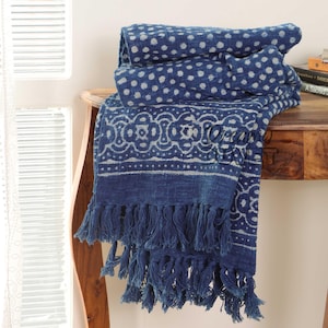 Puede incluir: Una manta de algodón azul con un patrón geométrico blanco y flecos. La manta está doblada y drapeada sobre una mesa de madera.