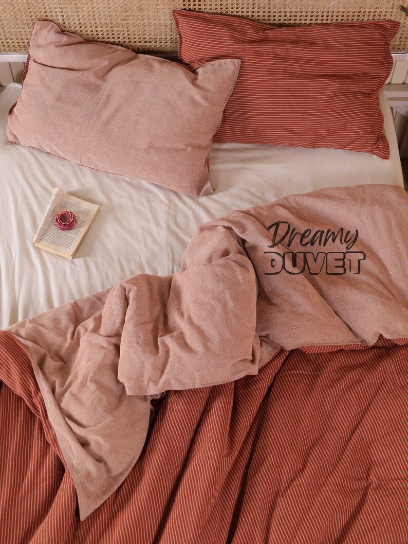 May include: A bed set featuring a light pink duvet cover and pillowcases. The duvet cover has the text "Dreamy DUVET". One pillowcase is plain pink, the other is striped. A book with a pink flower rests on the white sheet.
