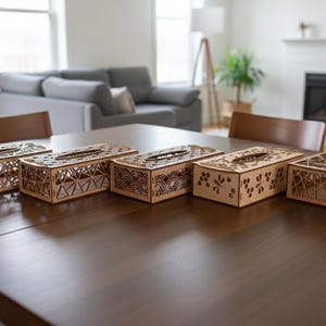以下が含まれることがあります： 幾何学模様や花柄など、それぞれ異なるレーザーカットデザインが施された、木製のティッシュボックスカバー5つ。明るい色の木材で作られており、濃い茶色のテーブルの上に置かれています。