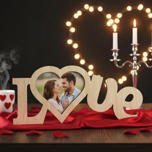 May include: A wooden "Love" photo frame with a heart-shaped cutout holding a couple's photo. A white mug with red hearts sits to the left, and a candelabra with lit candles is in the background. Red fabric and rose petals complete the romantic scene.