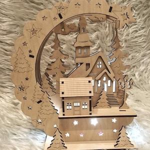 Puede incluir: Una escena de pueblo navide&ntilde;o de madera con una iglesia, casas y &aacute;rboles, iluminada por peque&ntilde;as luces. El dise&ntilde;o incluye un arco festoneado con estrellas y una estrella fugaz. La pieza est&aacute; hecha de madera clara.