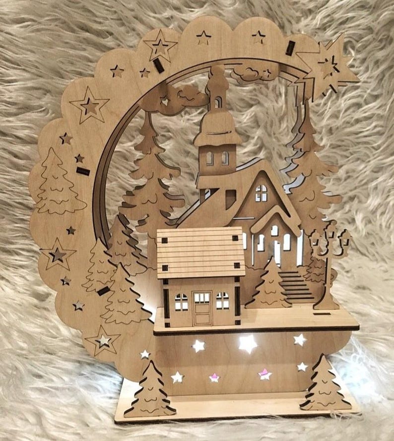Puede incluir: Una escena de pueblo navide&ntilde;o de madera con una iglesia, casas y &aacute;rboles, iluminada por peque&ntilde;as luces. El dise&ntilde;o incluye un arco festoneado con estrellas y una estrella fugaz. La pieza est&aacute; hecha de madera clara.