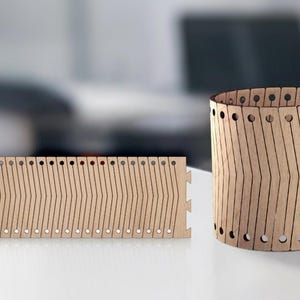 May include: Two wooden laser-cut pieces. One is a flat, rectangular panel with a series of vertical lines and small holes. The other is a cylindrical shape, also made of wood, with similar lines and holes. Both are light brown.