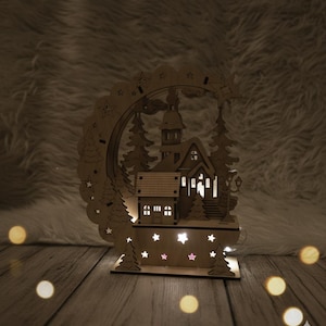 Puede incluir: Una escena de pueblo navide&ntilde;o de madera con una iglesia, &aacute;rboles y un arco decorativo. La escena est&aacute; iluminada con luces blancas c&aacute;lidas, creando un ambiente acogedor. La base presenta recortes en forma de estrella.