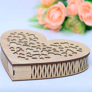 May include: A heart-shaped wooden box with a decorative cutout design on the lid. The box is made of light-coloured wood and has a detailed, geometric pattern around the sides. The background features peach-coloured flowers.