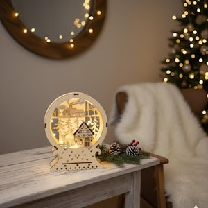 Puede incluir: Decoraci&oacute;n navide&ntilde;a de madera con una escena invernal con una casa, &aacute;rboles y un ciervo, iluminada por luces c&aacute;lidas. El dise&ntilde;o redondo se asienta sobre una base de madera, creando una exhibici&oacute;n festiva.