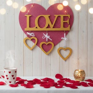 May include: A Valentine's Day themed display featuring a large pink glitter heart with the word "LOVE" in gold letters. Three hanging gold heart ornaments and a white mug with red hearts are visible. Red rose petals and a candle holder complete the scene.