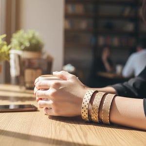 May include: A person holding a white coffee cup, wearing three gold-colored bracelets with different patterns. The bracelets are on the person's wrist. A smartphone rests on a wooden table, with plants and a window in the background.