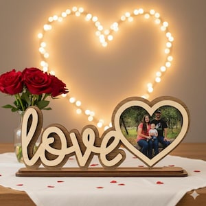 May include: A wooden photo frame in the shape of a heart, holding a family photo, sits on a table next to a wooden "love" sign. Red roses in a glass vase and a heart-shaped light display are in the background.