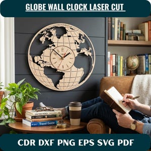 May include: A circular laser-cut wooden wall clock featuring a world map design with a central clock face displaying black numerals and hands. The clock is mounted on a dark horizontal paneled wall above a wooden table with books and a cup.