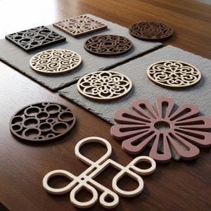Op de afbeelding: Een verzameling decoratieve onderzetters in verschillende vormen en kleuren. De onderzetters hebben vierkante, ronde en bloemvormige ontwerpen, met ingewikkelde uitgesneden patronen. Kleuren zijn onder meer zwart, bruin, beige en mauve. Ze zijn gerangschikt op een donkere houten tafel.