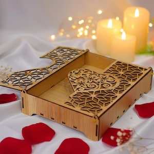 以下が含まれることがあります： ハート型の切り抜きと複雑な幾何学模様が施された木製の箱。箱は開いており、内部が見えます。赤いバラの花びらが、白いサテンの表面に箱の周りに散らばっています。背景には、ろうそくの灯りと花があります。