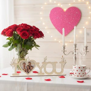 May include: A romantic scene featuring a bouquet of red roses in a glass vase, a wooden "I LOVE YOU" photo frame, a candelabra with three white candles, and a heart-decorated teacup. Red rose petals are scattered on the white tablecloth.