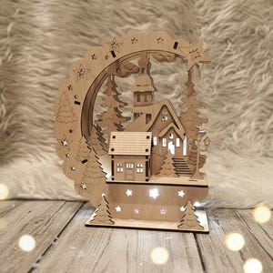 Puede incluir: Una escena de pueblo navide&ntilde;o de madera con una iglesia, casas y &aacute;rboles. La escena est&aacute; iluminada con peque&ntilde;as luces, y una forma de estrella recortada est&aacute; en la parte superior. La estructura de madera est&aacute; sobre una base de madera.